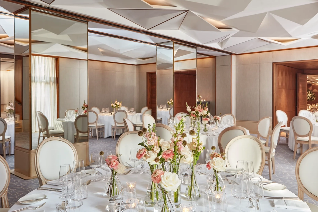 Round tables with white chairs, floral centrepieces, and mirrored walls reflecting soft daylight.