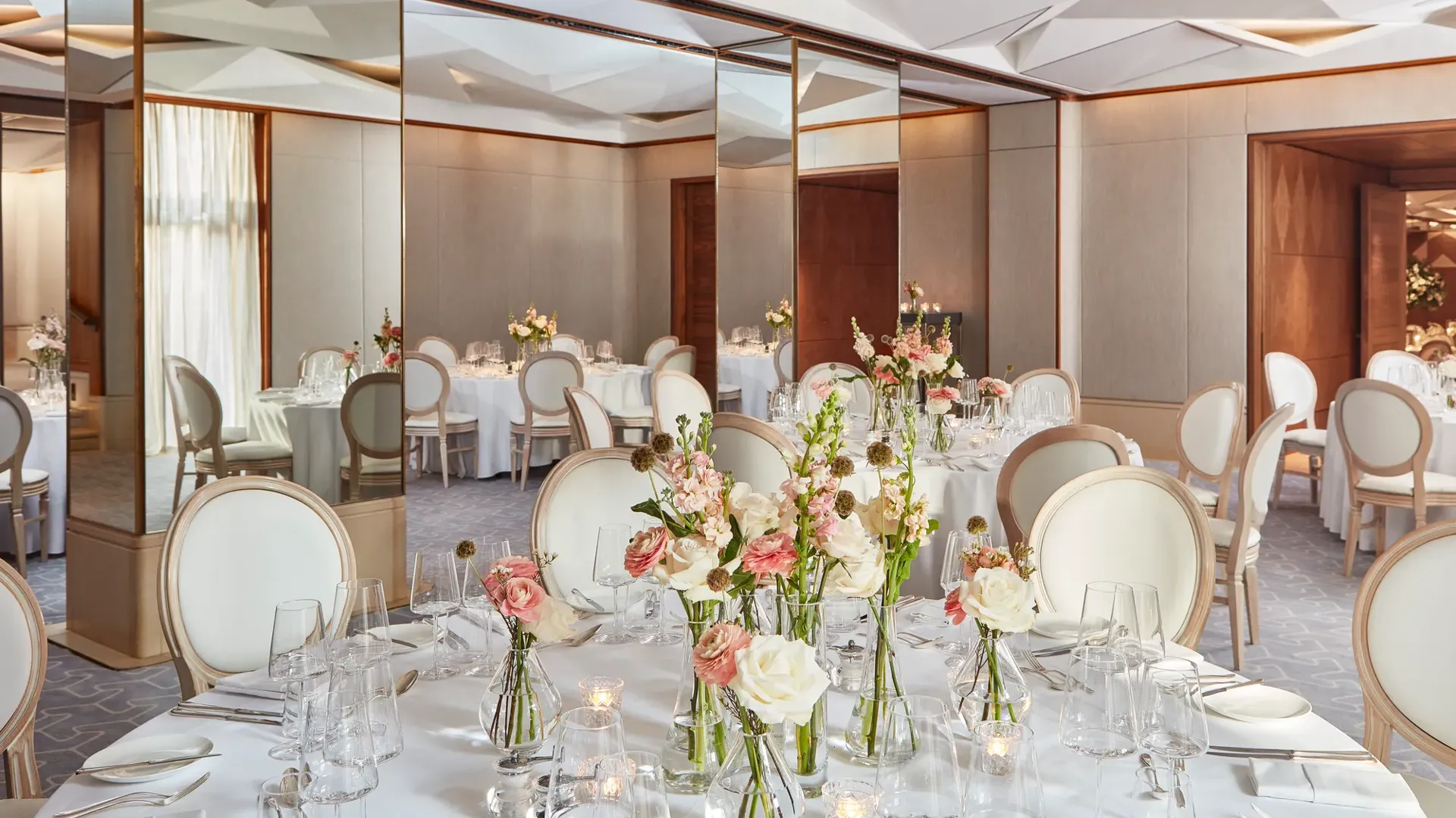 Round tables with white chairs, floral centrepieces, and mirrored walls reflecting soft daylight.