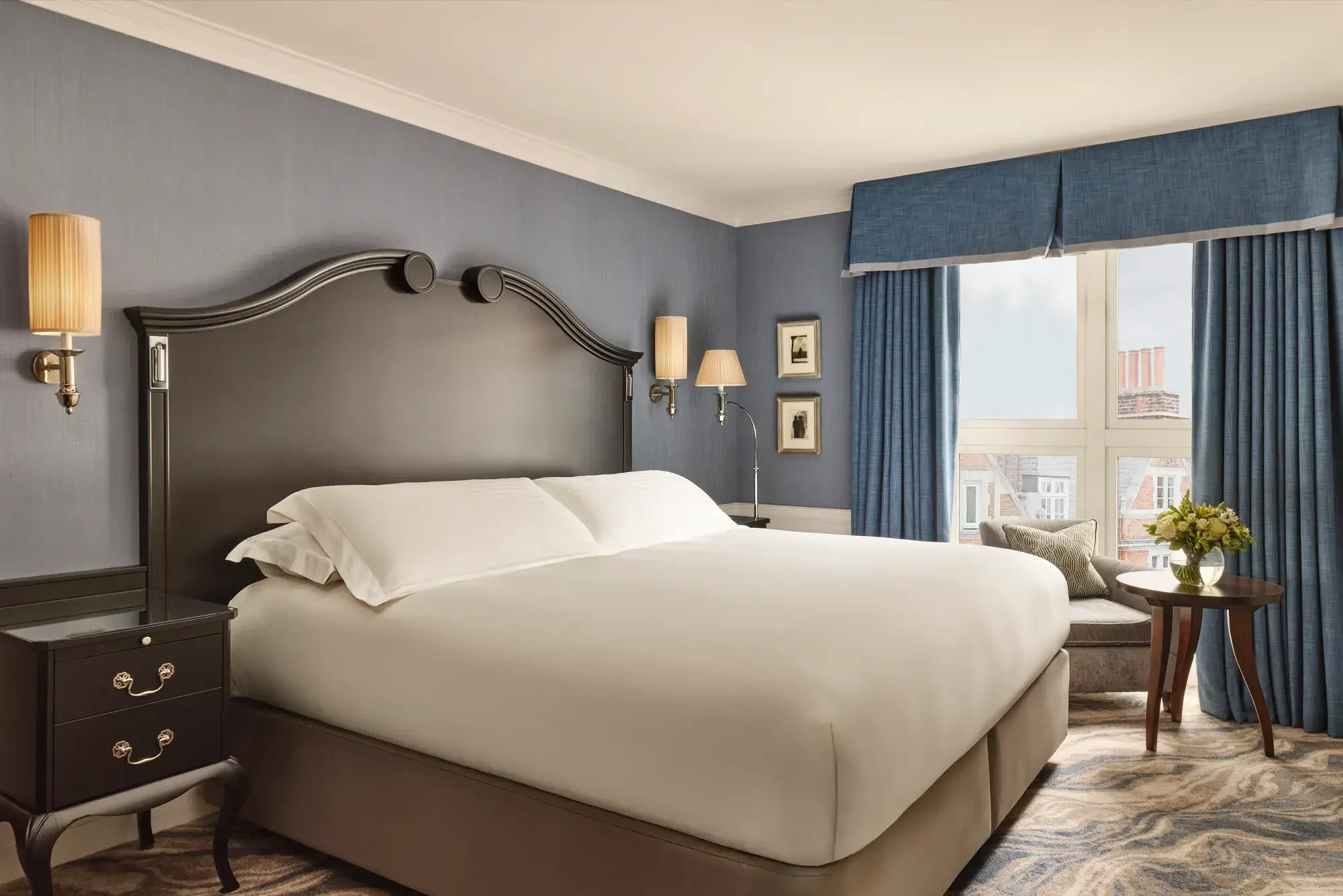Elegant bedroom with a large bed, dark wood headboard, blue walls and curtains, and a window overlooking rooftops.