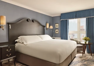 Elegant bedroom with a large bed, dark wood headboard, blue walls and curtains, and a window overlooking rooftops.