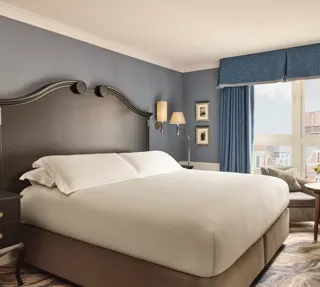 Elegant bedroom with a large bed, dark wood headboard, blue walls and curtains, and a window overlooking rooftops.