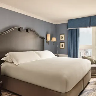 Elegant bedroom with a large bed, dark wood headboard, blue walls and curtains, and a window overlooking rooftops.