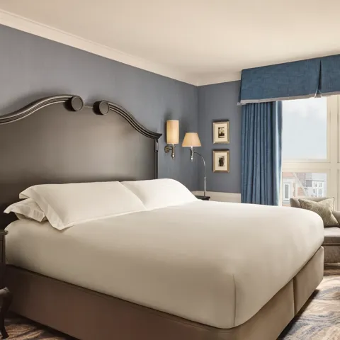Elegant bedroom with a large bed, dark wood headboard, blue walls and curtains, and a window overlooking rooftops.