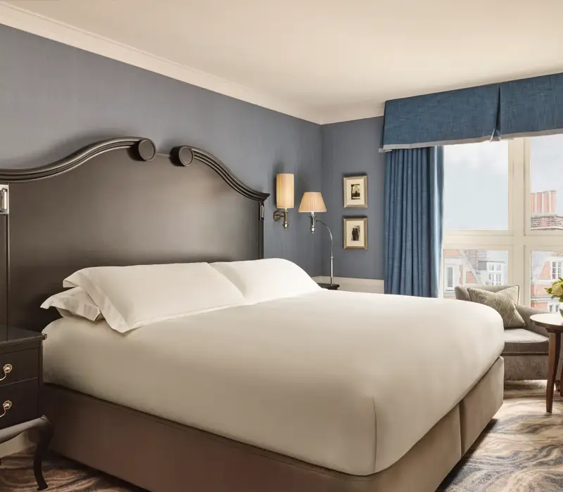 Elegant bedroom with a large bed, dark wood headboard, blue walls and curtains, and a window overlooking rooftops.