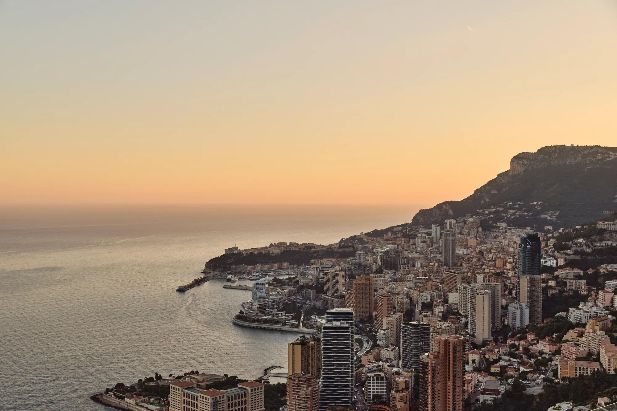 Monaco skyline at sunset with coastal high-rises, harbour, and mountains under warm golden light
