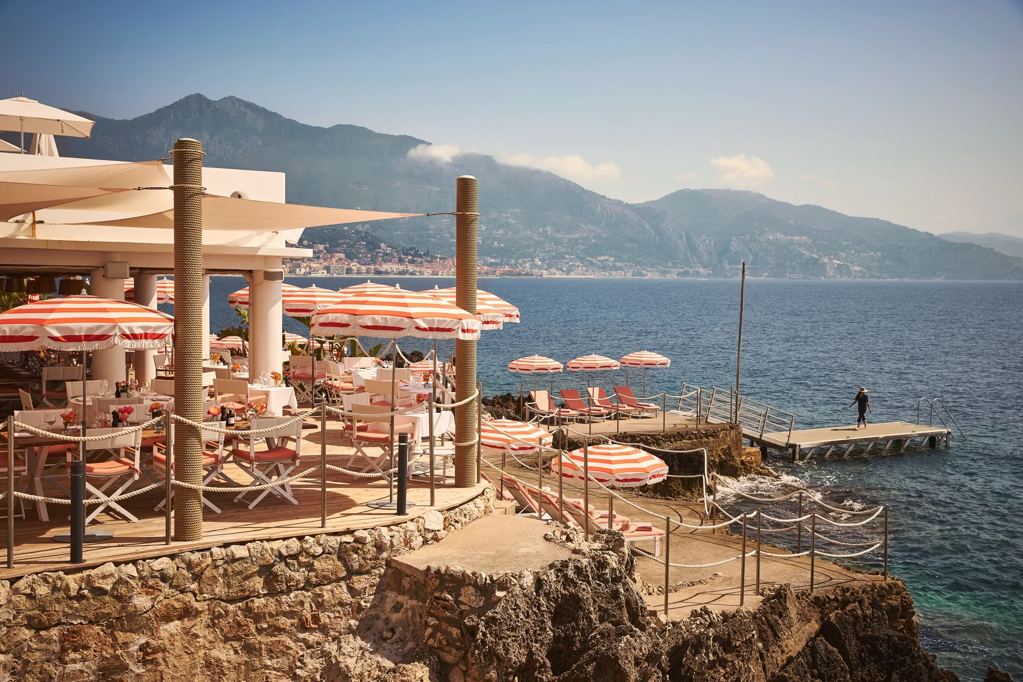 Seaside restaurant terrace with red-striped umbrellas and sun loungers on rocky shore, overlooking ocean and mountains