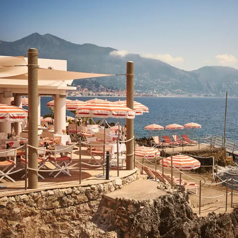 Seaside restaurant terrace with red-striped umbrellas and sun loungers on rocky shore, overlooking ocean and mountains