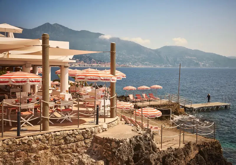 Seaside restaurant terrace with red-striped umbrellas and sun loungers on rocky shore, overlooking ocean and mountains