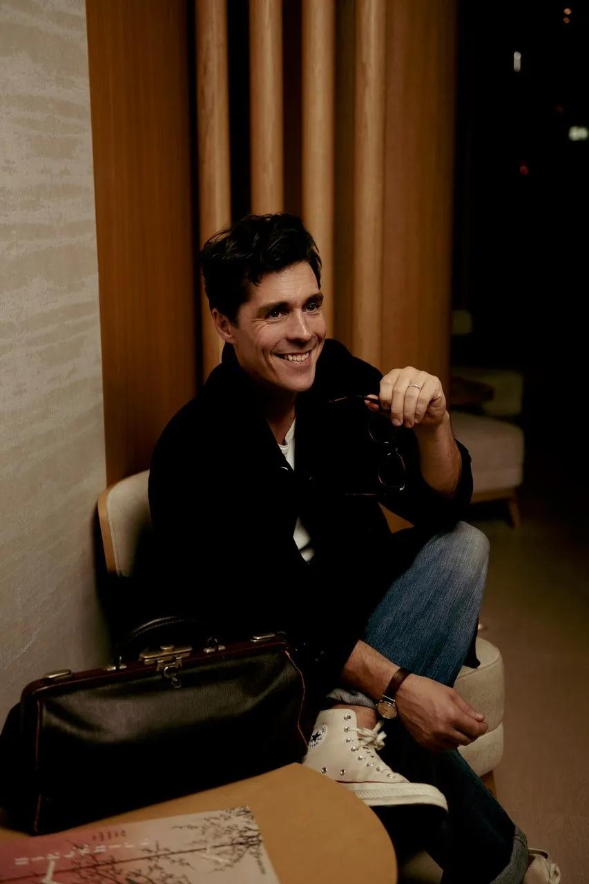 A man sitting in a lounge area at The Maybourne Riviera, smiling while holding a pair of glasses, with a leather bag resting on the table beside him.