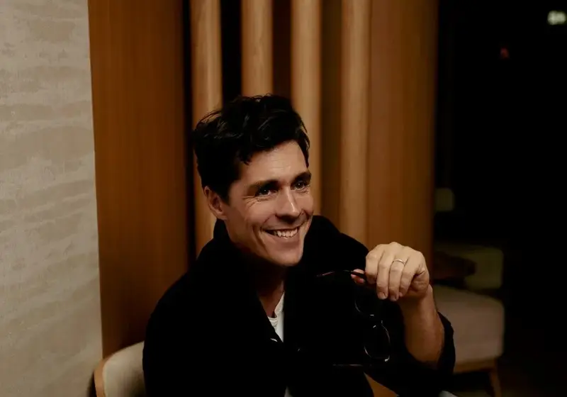 A man sitting in a lounge area at The Maybourne Riviera, smiling while holding a pair of glasses, with a leather bag resting on the table beside him.