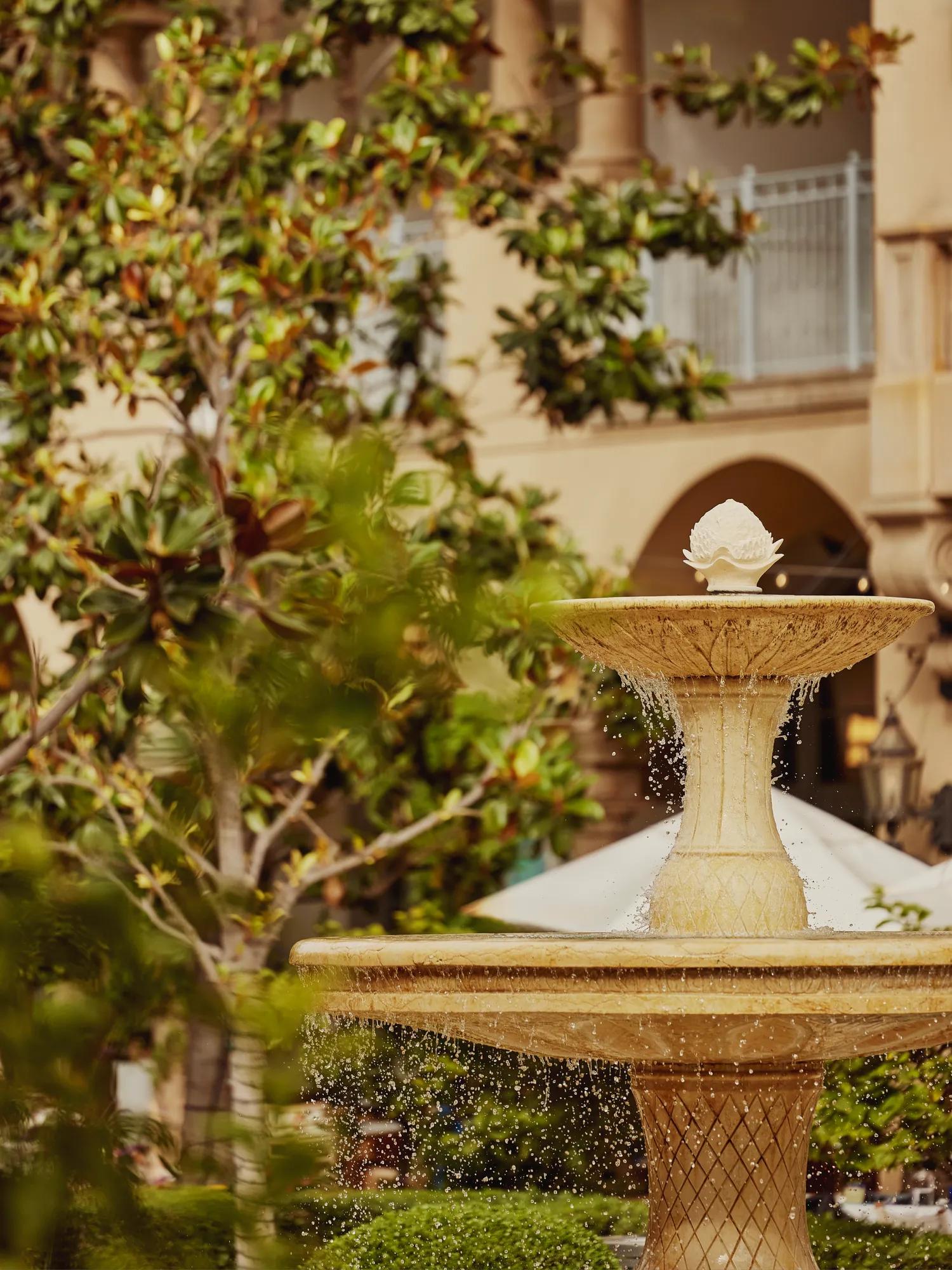 Tiered stone fountain splashing in verdant courtyard beside arched hotel colonnade.