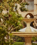 Tiered stone fountain splashing in verdant courtyard beside arched hotel colonnade.