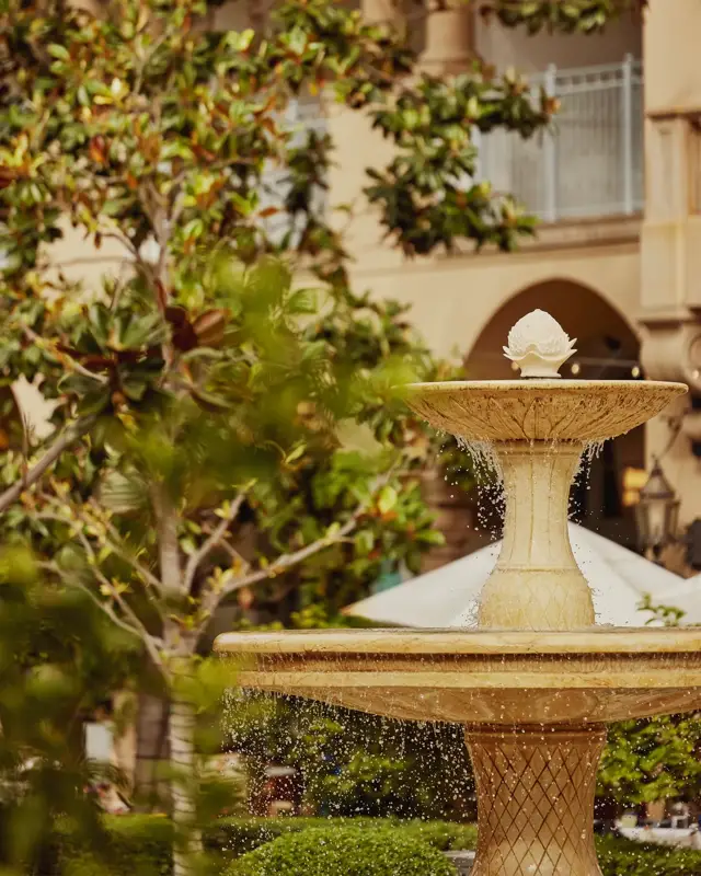 Tiered stone fountain splashing in verdant courtyard beside arched hotel colonnade.