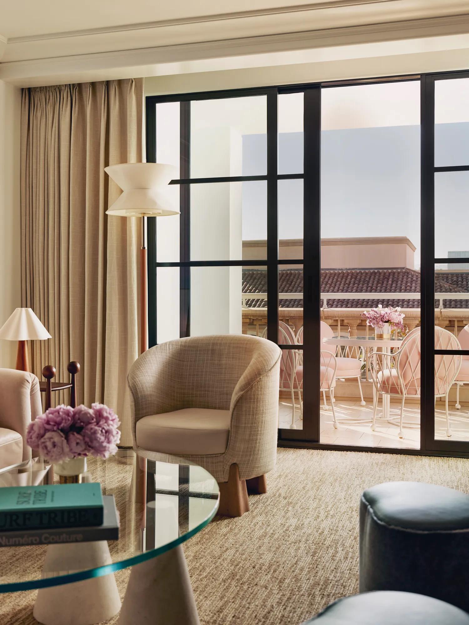 Sitting area with armchairs and a glass coffee table opening to a terrace furnished with pink chairs.