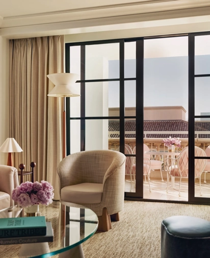 Sitting area with armchairs and a glass coffee table opening to a terrace furnished with pink chairs.