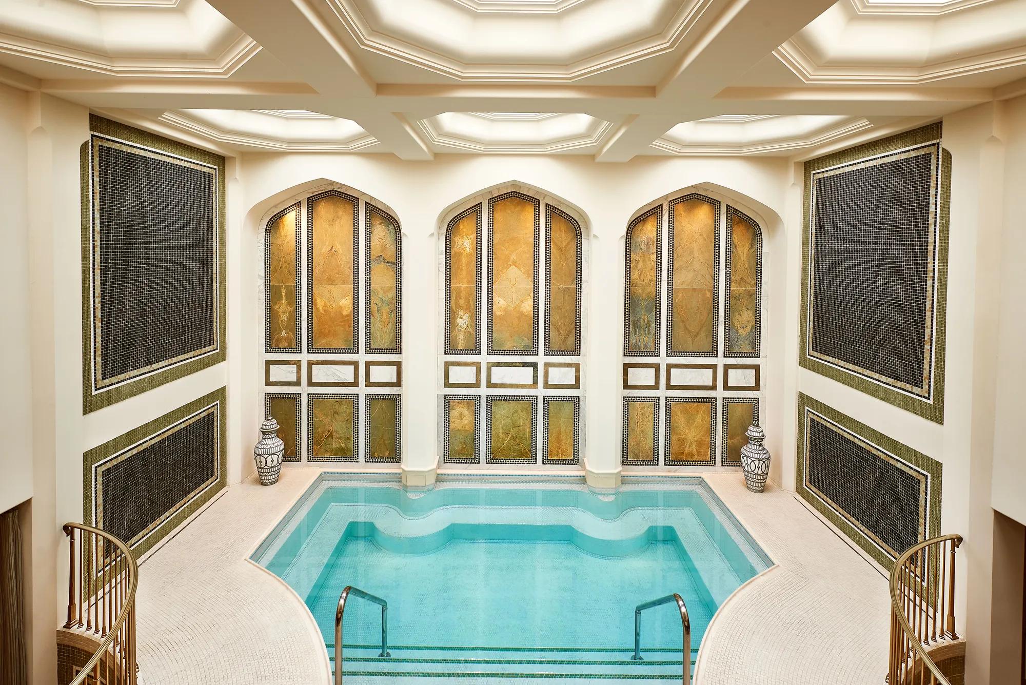 **Alt text:**  
Elegant indoor mineral pool surrounded by cream-colored walls with intricate mosaic tile panels and three arched insets featuring gold-toned stone. The rectangular pool has softly curved edges and shallow steps with metal handrails. Decorative ceramic vases sit on either side, and a coffered ceiling with recessed lighting creates a warm, spa-like atmosphere.