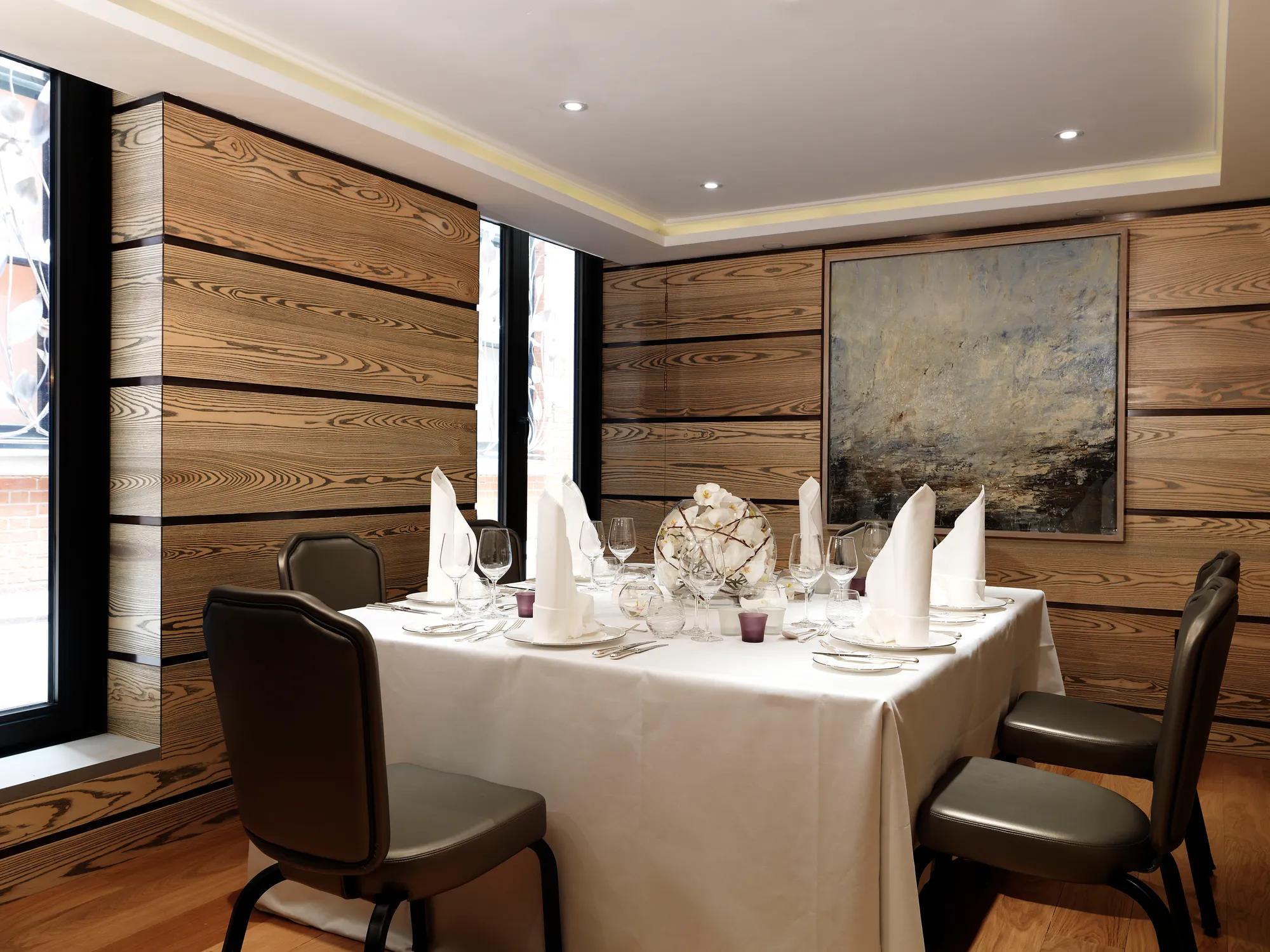 Private dining room with a square table dressed in white linens, folded napkins and glassware, surrounded by wood-panelled walls and contemporary artwork.
