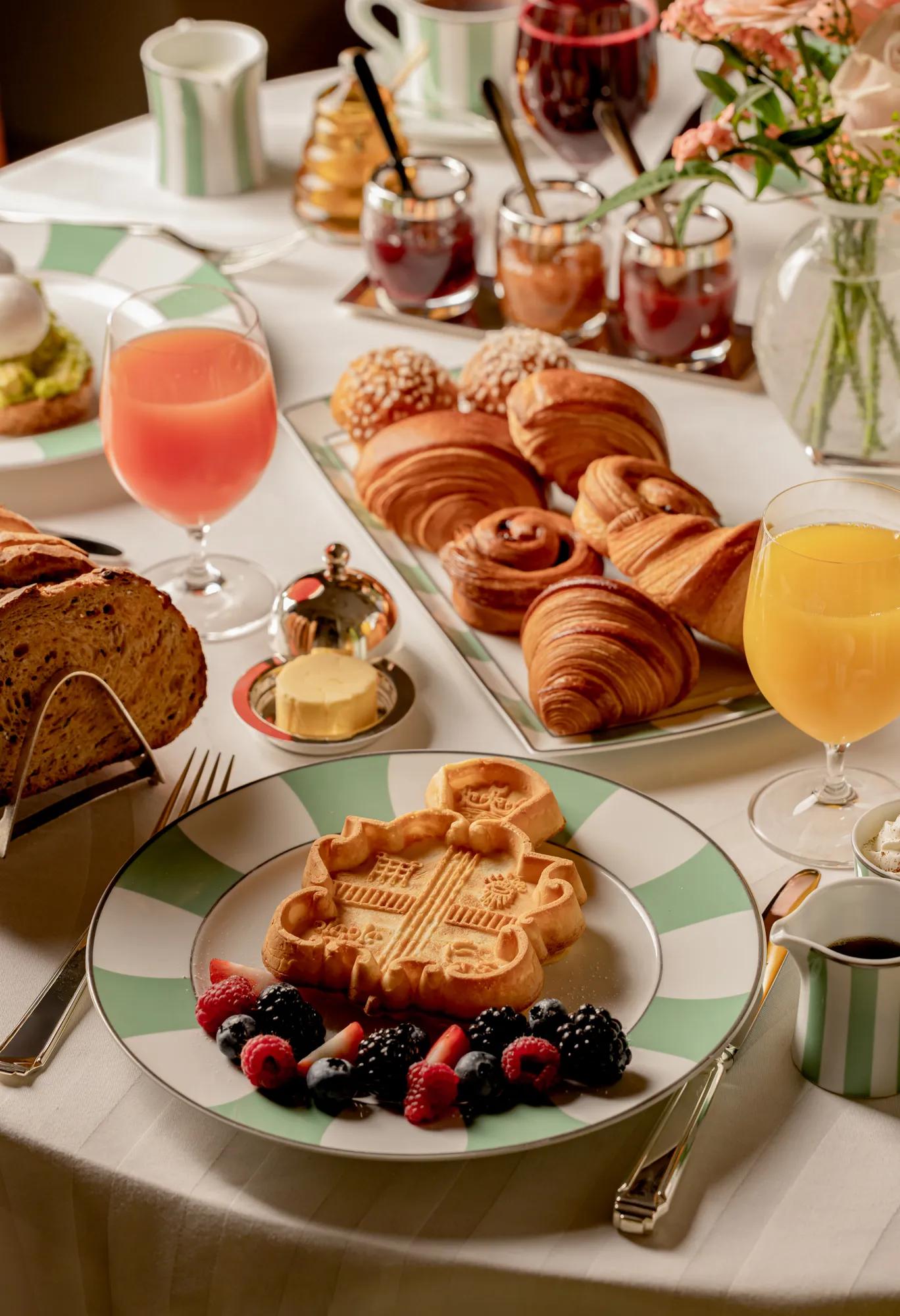 Breakfast spread with waffles, croissants, pastries, fruit, bread, and juices arranged on a white tablecloth.