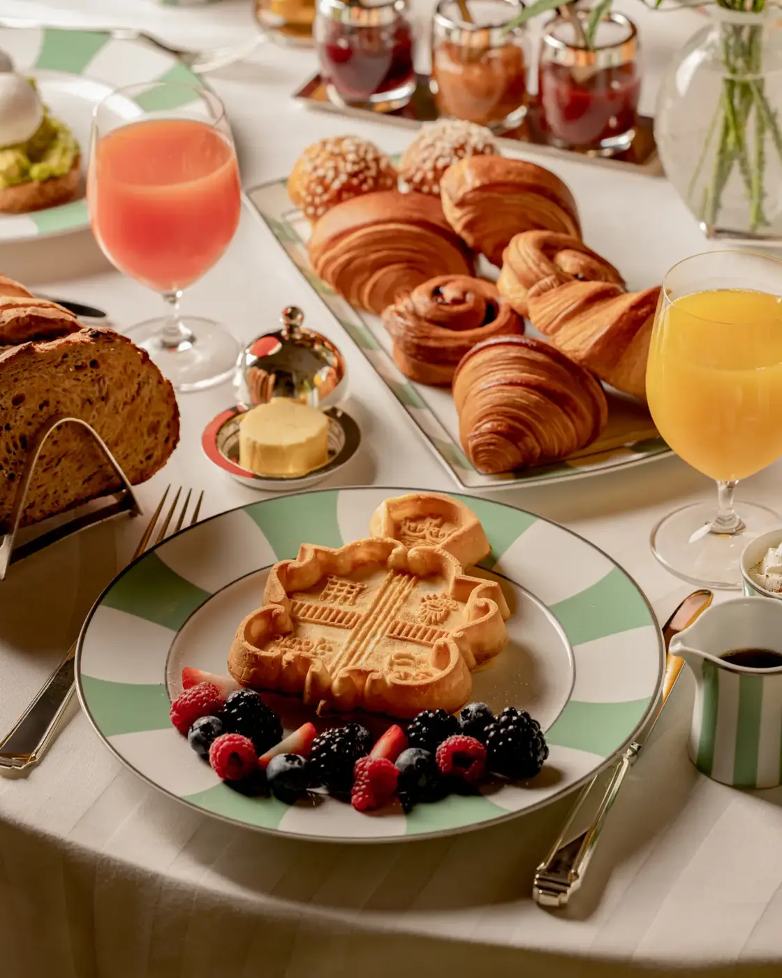 Breakfast spread with waffles, croissants, pastries, fruit, bread, and juices arranged on a white tablecloth.