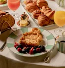 Breakfast spread with waffles, croissants, pastries, fruit, bread, and juices arranged on a white tablecloth.