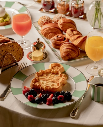Breakfast spread with waffles, croissants, pastries, fruit, bread, and juices arranged on a white tablecloth.