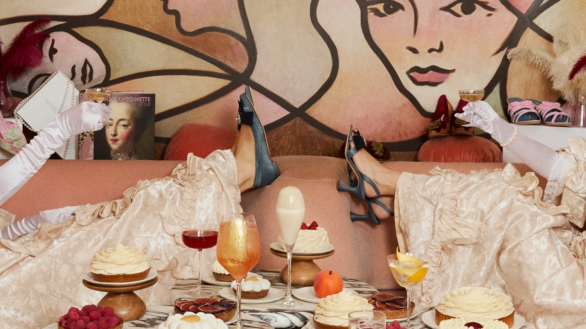 Two women lounging on a sofa in vintage-style dresses and heels, enjoying desserts and cocktails amid playful, artistic décor.