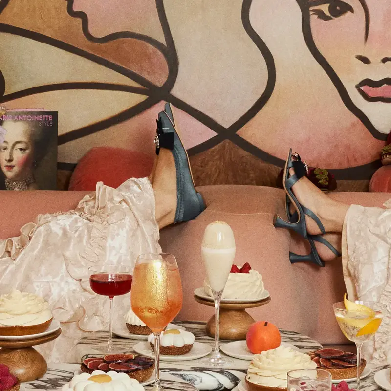 Two women lounging on a sofa in vintage-style dresses and heels, enjoying desserts and cocktails amid playful, artistic décor.