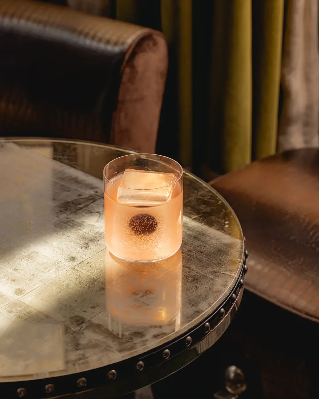 A pale cocktail with a large ice cube on a reflective metal table beside leather seating and velvet curtains.