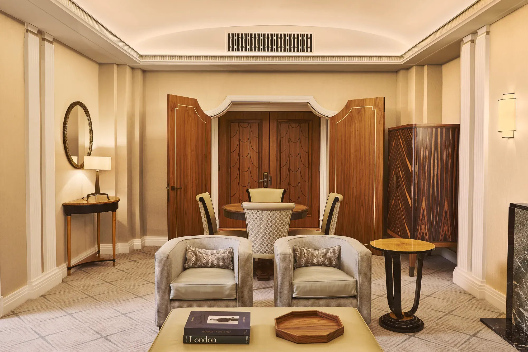 Elegant suite living area with two armchairs, coffee table, round dining table, wood-panelled doors and warm lighting.