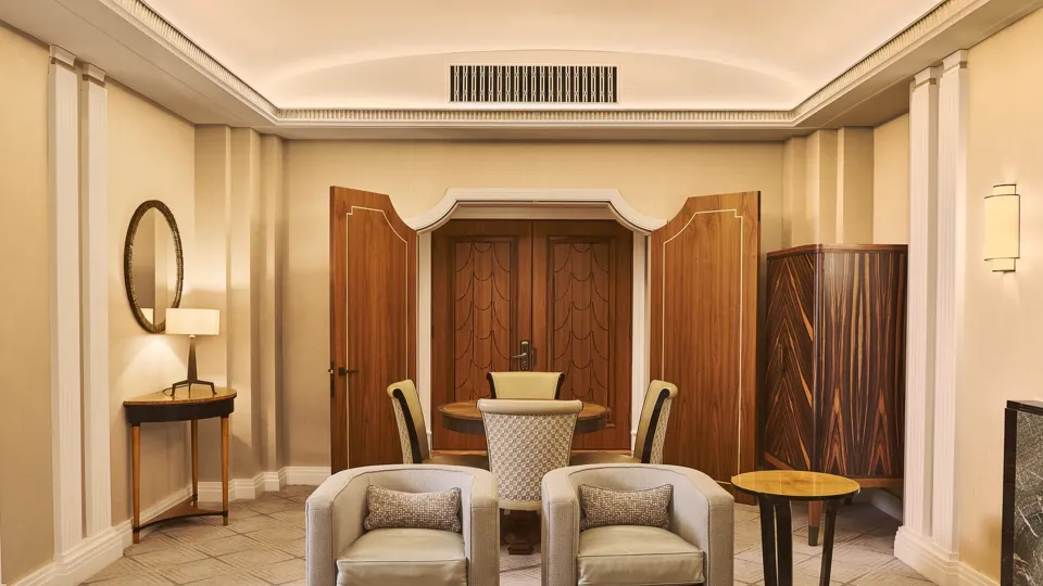 Elegant suite living area with two armchairs, coffee table, round dining table, wood-panelled doors and warm lighting.