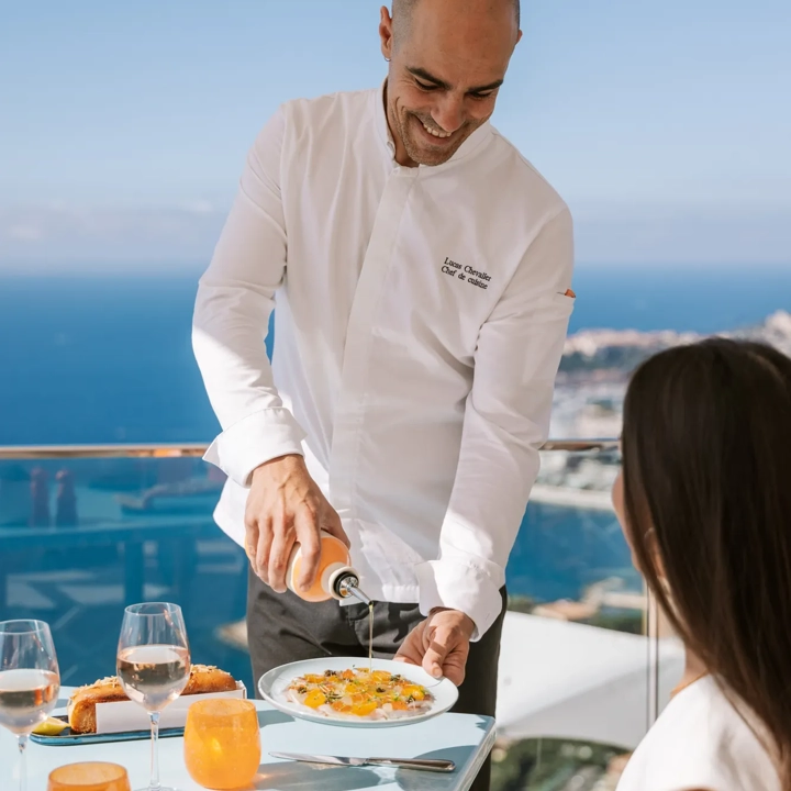 Un serveur verse une sauce sur le plat d’une cliente sur une terrasse ensoleillée face à la mer.
