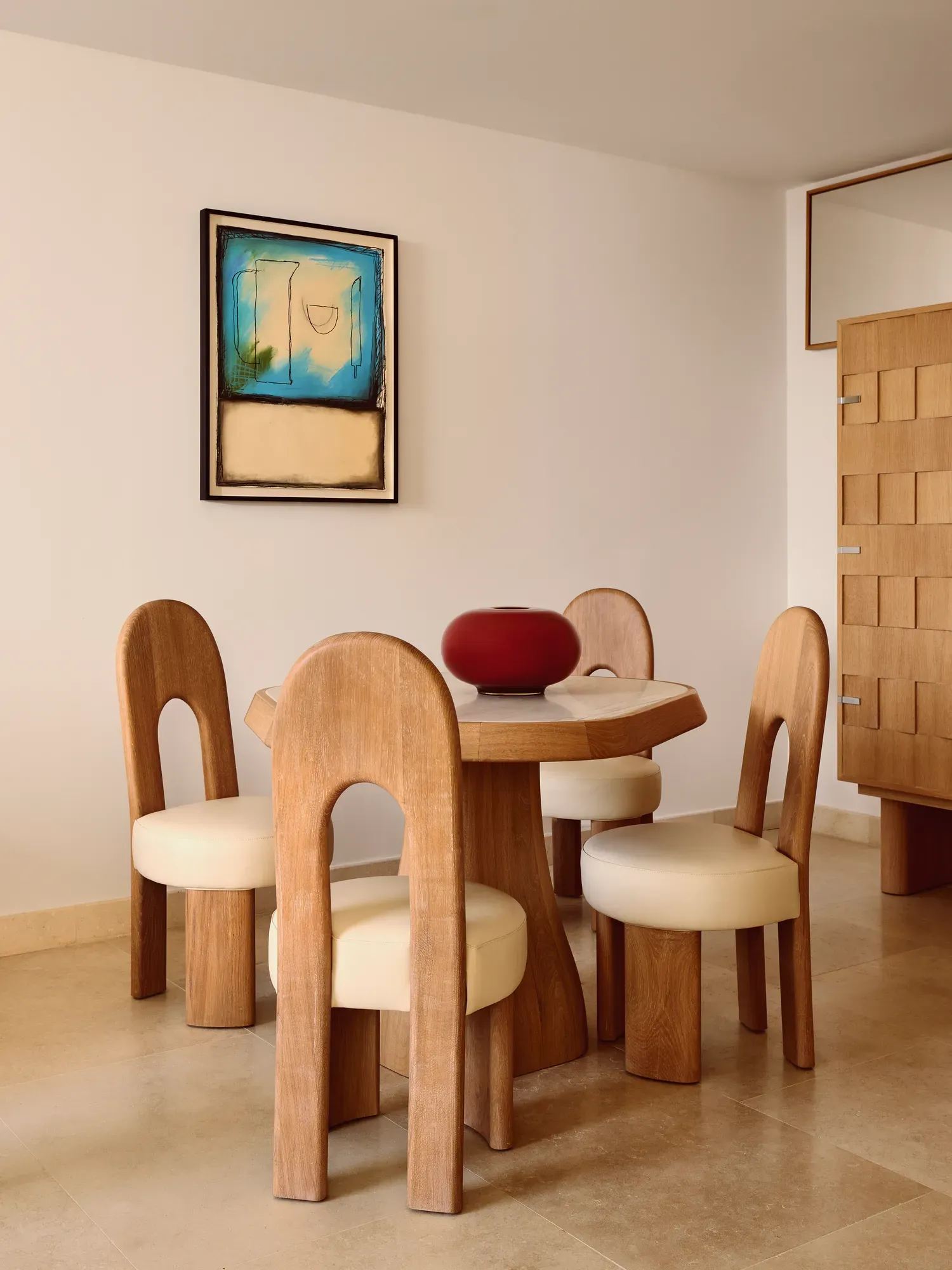 Contemporary dining area with a round wooden table, four sculptural chairs with cream cushions, a bold red vase centerpiece, and abstract artwork on the wall.