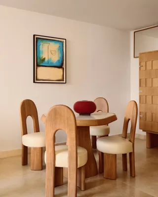 Contemporary dining area with a round wooden table, four sculptural chairs with cream cushions, a bold red vase centerpiece, and abstract artwork on the wall.