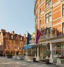 Exterior of The Connaught hotel in Mayfair, London, showing a curved red-brick façade with cream stone detailing, three flags over the entrance canopy, and potted trees by the doorway, with adjacent period buildings under a clear sky.