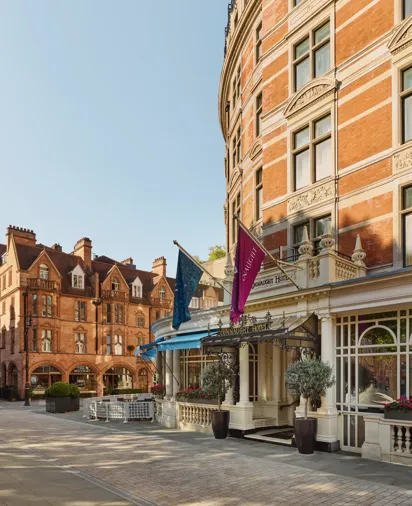 Exterior of The Connaught hotel in Mayfair, London, showing a curved red-brick façade with cream stone detailing, three flags over the entrance canopy, and potted trees by the doorway, with adjacent period buildings under a clear sky.