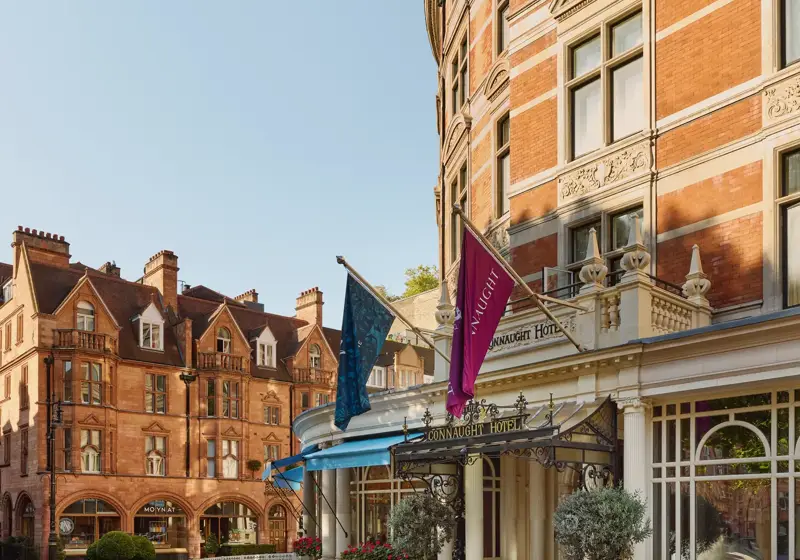 Exterior of The Connaught hotel in Mayfair, London, showing a curved red-brick façade with cream stone detailing, three flags over the entrance canopy, and potted trees by the doorway, with adjacent period buildings under a clear sky.