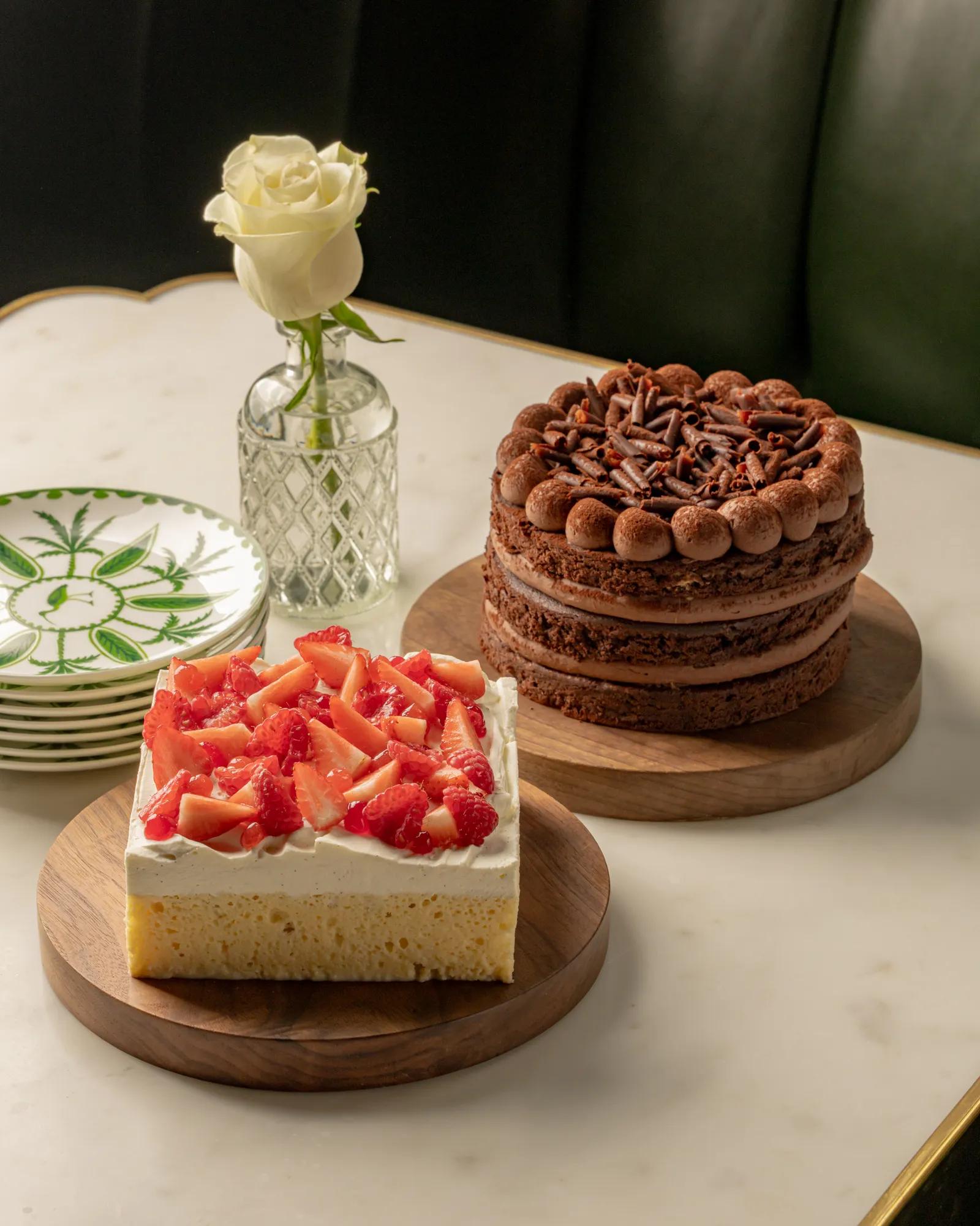 Strawberry-topped sponge cake and chocolate layer cake displayed on wooden boards beside a white rose in a glass vase on a marble table.