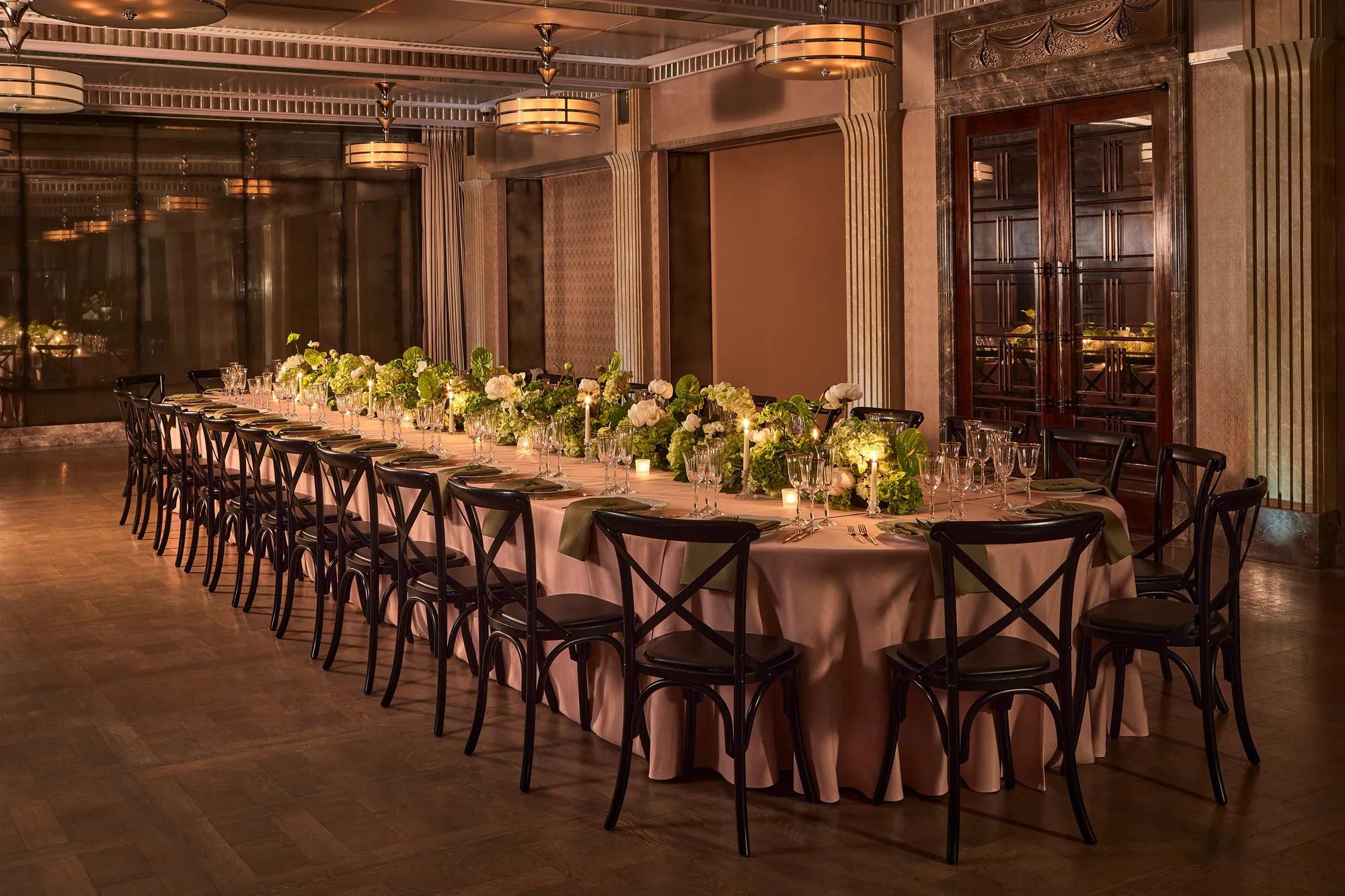Long banquet table with black chairs, green and white floral arrangements, and candlelight in an elegant dining room.