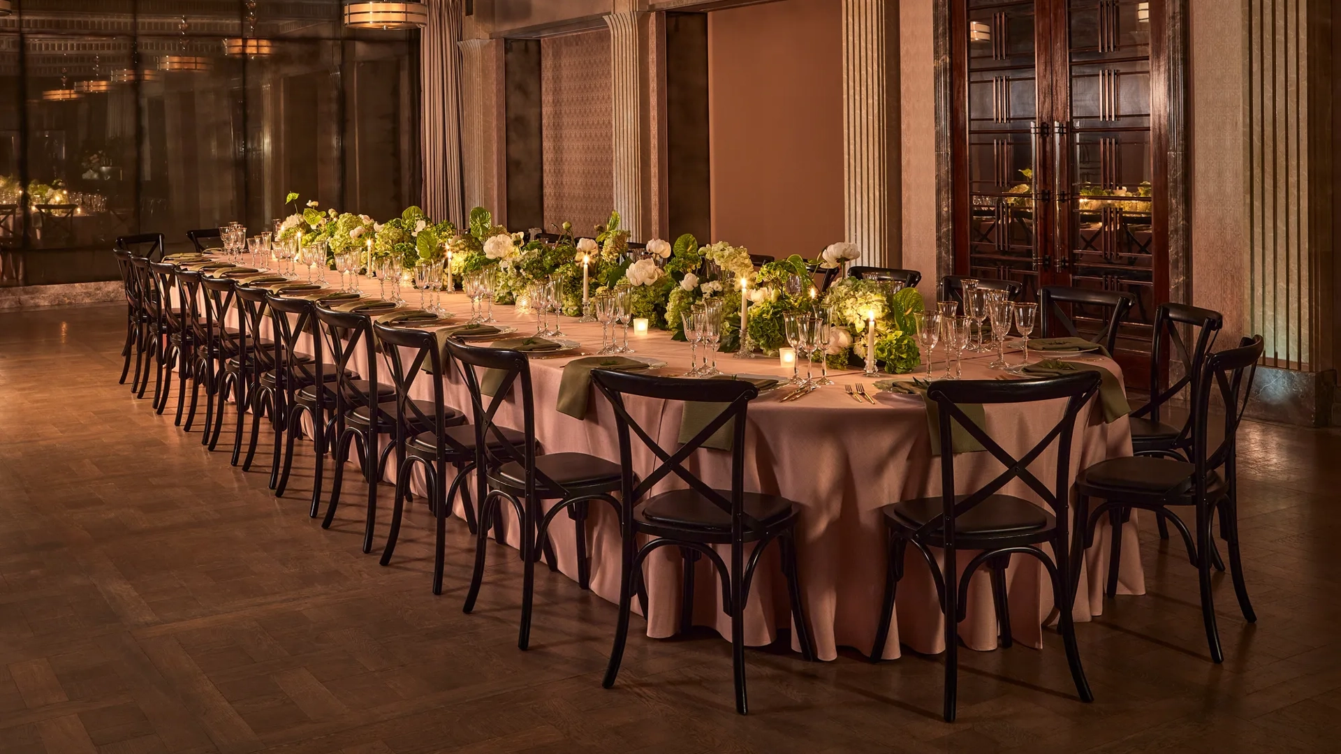 Long banquet table with black chairs, green and white floral arrangements, and candlelight in an elegant dining room.
