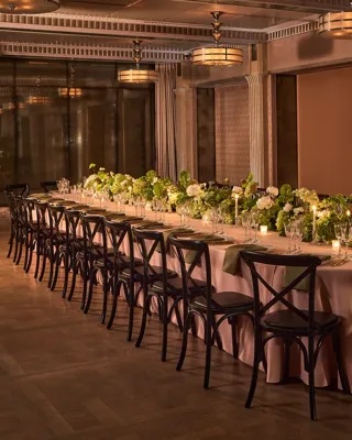Long banquet table with black chairs, green and white floral arrangements, and candlelight in an elegant dining room.