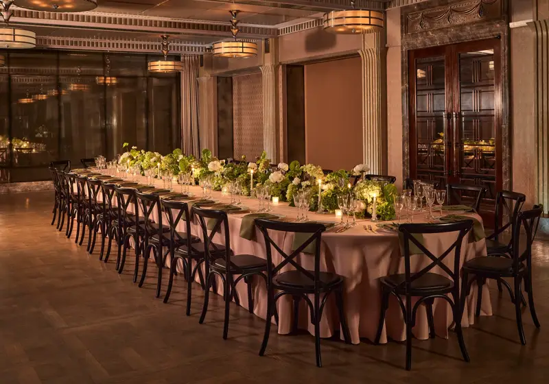 Long banquet table with black chairs, green and white floral arrangements, and candlelight in an elegant dining room.