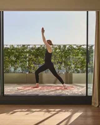 Person practicing yoga on a balcony, framed by curtains, with greenery and sunlight beyond glass doors.