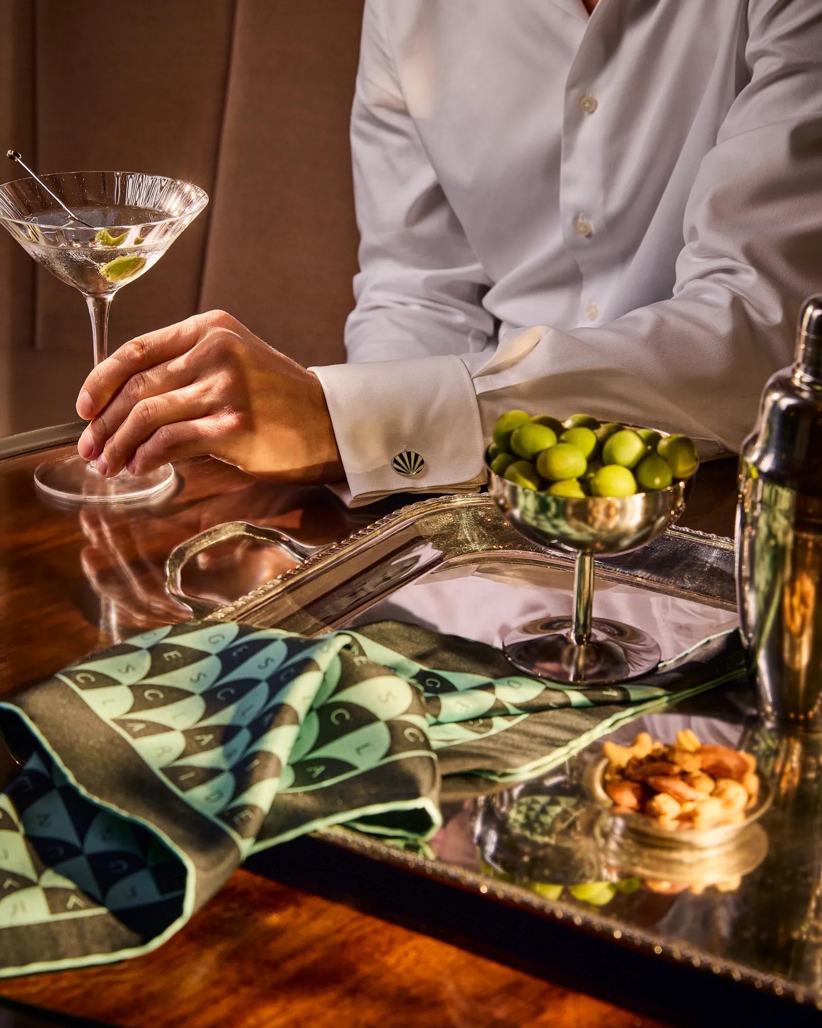 Guest holding a martini at a polished table with olives, bar snacks, a shaker, and patterned napkin arranged on a tray.