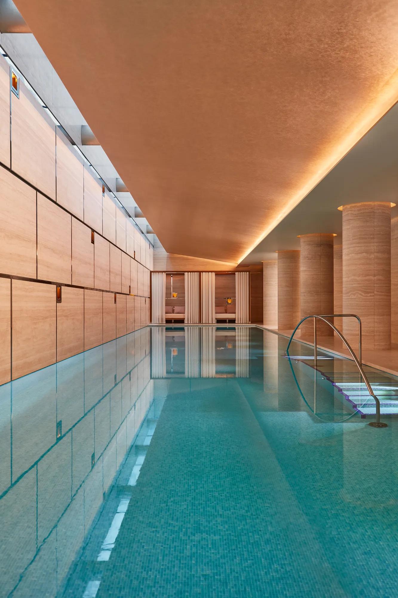 Indoor swimming pool with turquoise tiles, warm lighting, and stone columns, featuring metal handrails and calm reflective water beneath a softly illuminated ceiling.