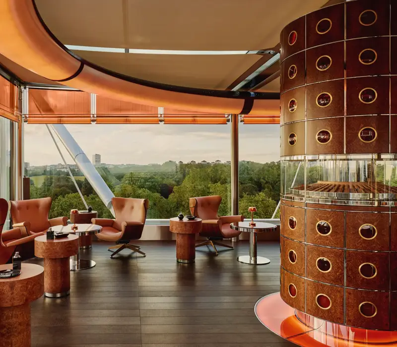 Cigar lounge with caramel leather chairs, circular tables, and a round humidor overlooking Hyde Park through floor-to-ceiling windows.