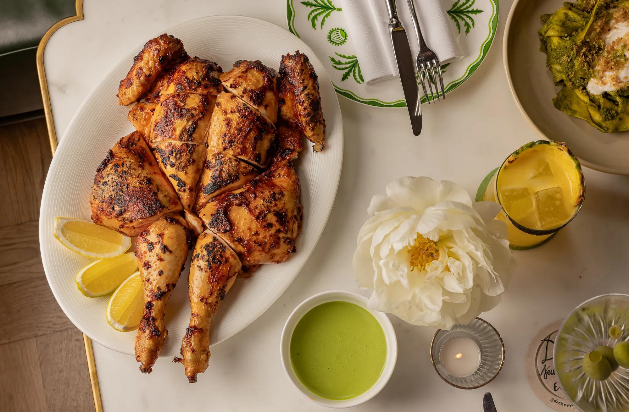 Whole grilled chicken on white platter with lemon wedges, green sauce, cocktail, and white flower on table.