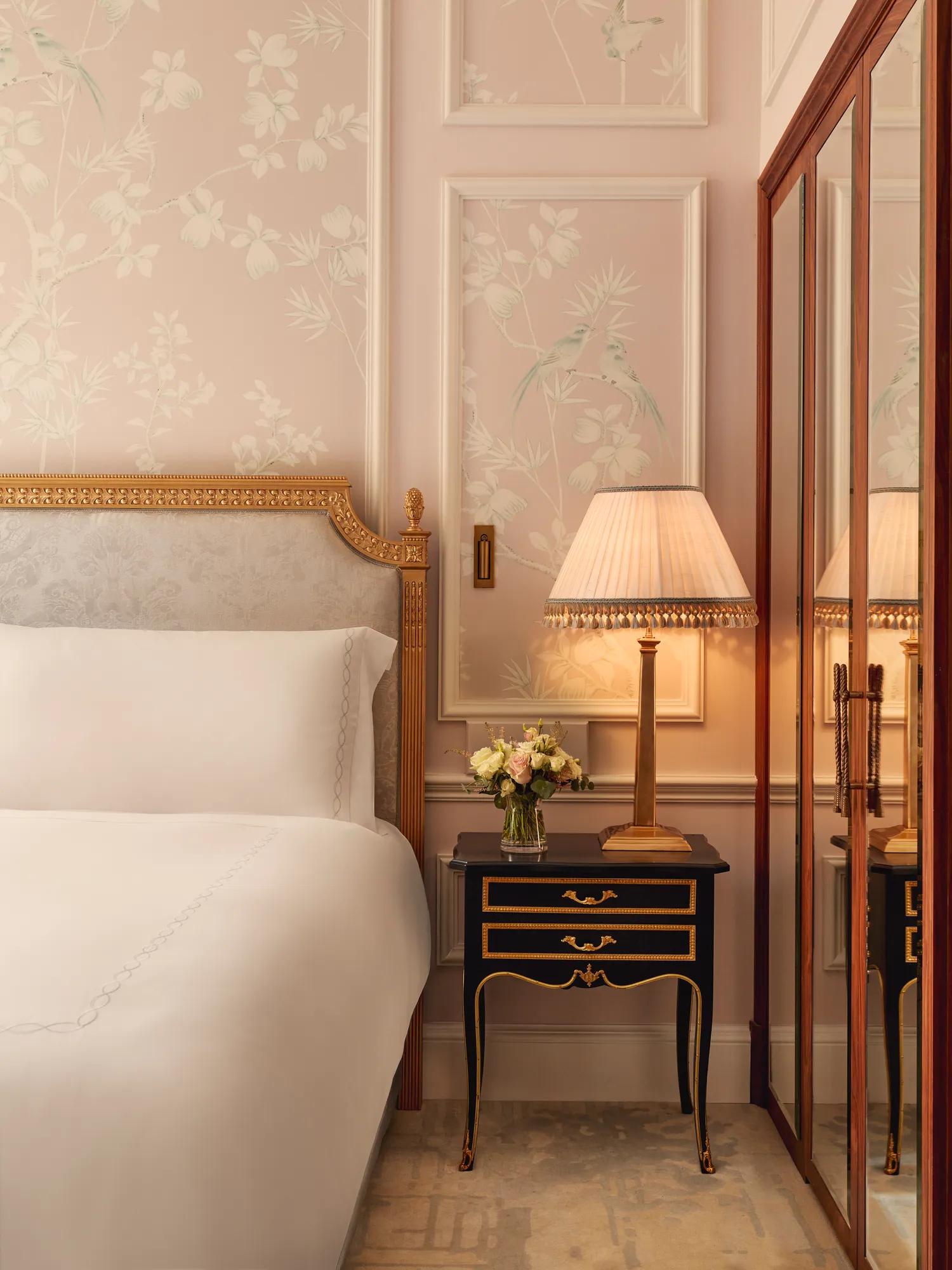 Elegant bedroom corner with upholstered bed, floral wallpaper, bedside table with lamp and flowers, and mirrored wardrobe reflecting the scene.