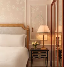 Elegant bedroom corner with upholstered bed, floral wallpaper, bedside table with lamp and flowers, and mirrored wardrobe reflecting the scene.