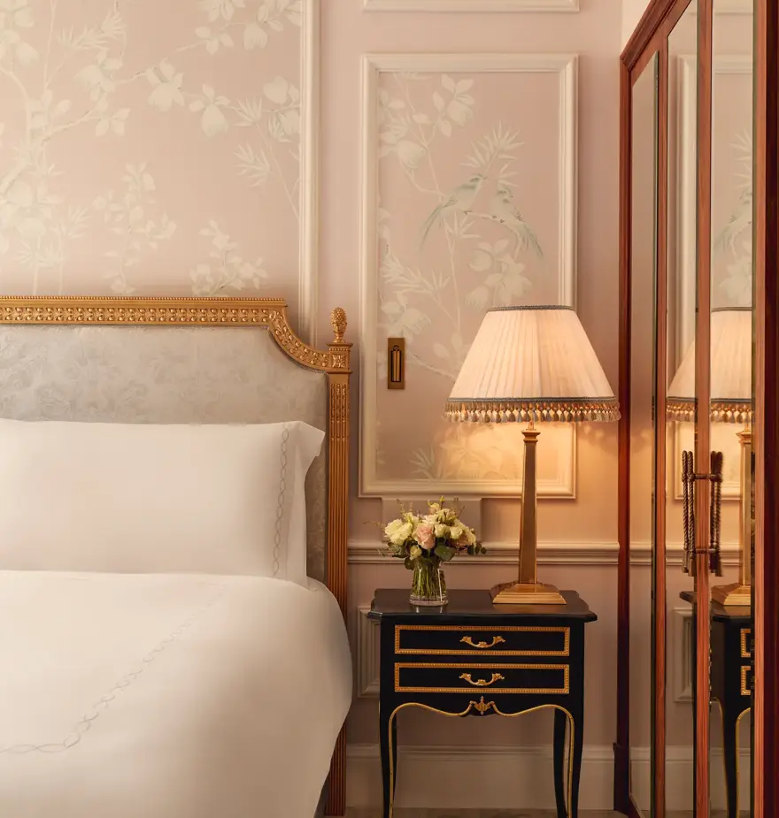 Elegant bedroom corner with upholstered bed, floral wallpaper, bedside table with lamp and flowers, and mirrored wardrobe reflecting the scene.