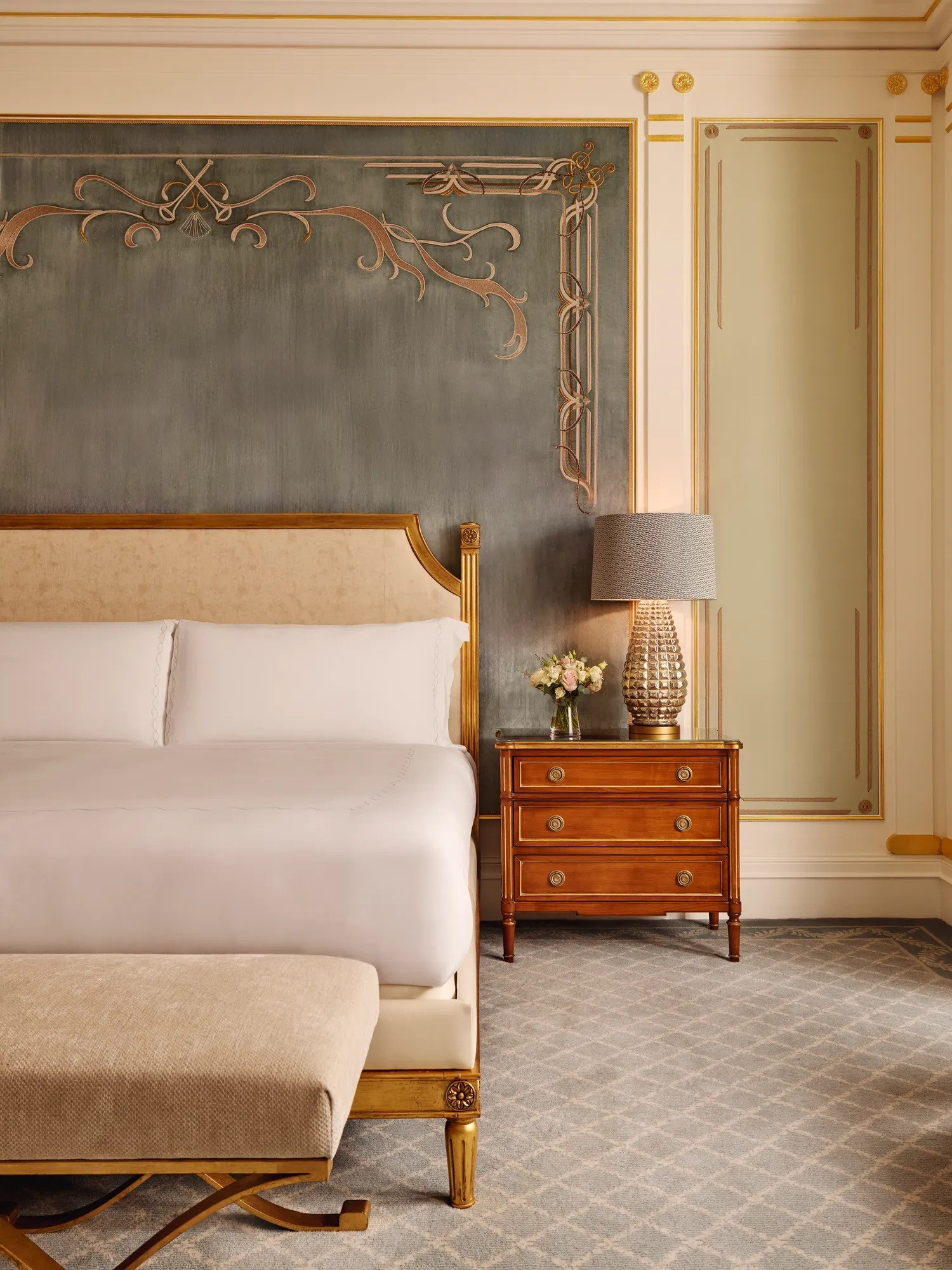Refined bedroom with a king bed featuring a gold-trimmed headboard, a wooden bedside table with a textured lamp and flowers, and an ornate wall with soft blue and gold detailing.
